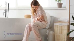 A woman sitting on a toilet, holding her lower abdomen in pain.